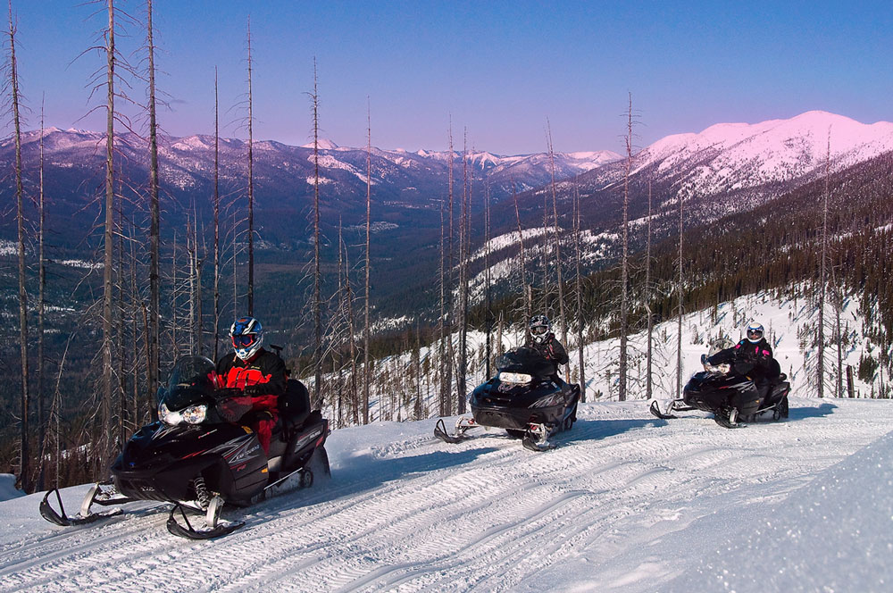 Three snowmobilers riding along a snowy mountain trail at sunset.