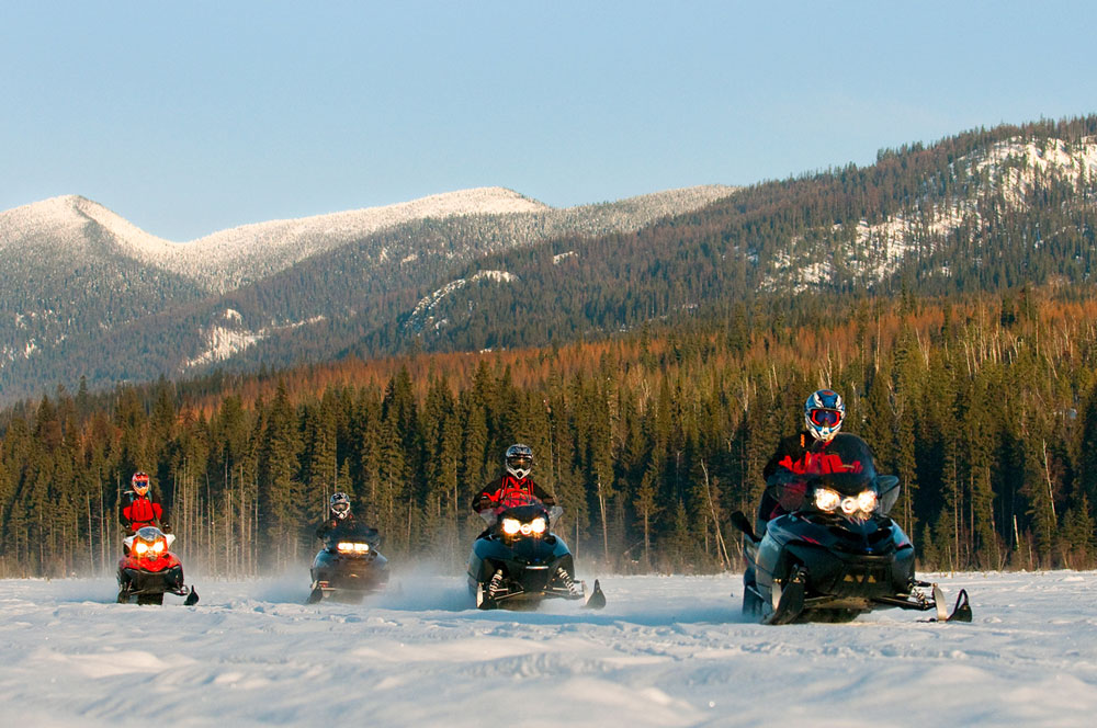 Four snowmobilers driving in formation through a scenic winter landscape.