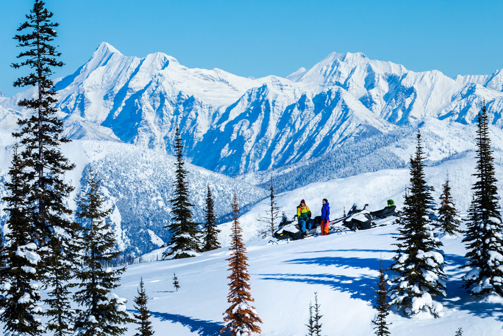 Group of snowmobilers resting on a snowy mountain ridge with towering peaks in the background.