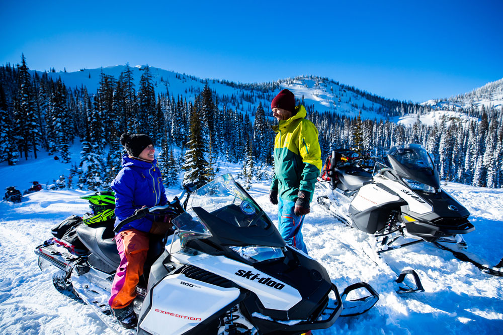 Two snowmobilers chatting during a break in a snowy mountain setting.